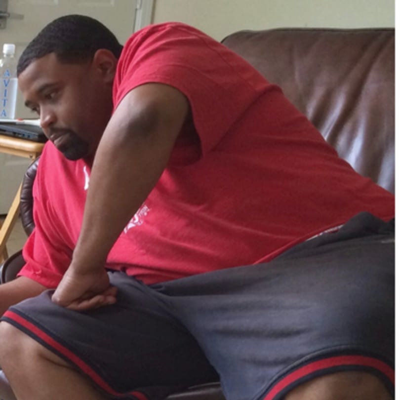 Man in a red shirt and dark shorts sitting on a brown leather couch, looking down.