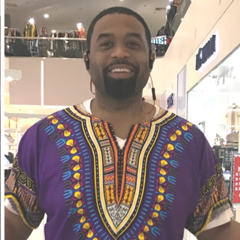 Smiling man in a purple dashiki shirt with earbuds, standing in a brightly lit indoor space.